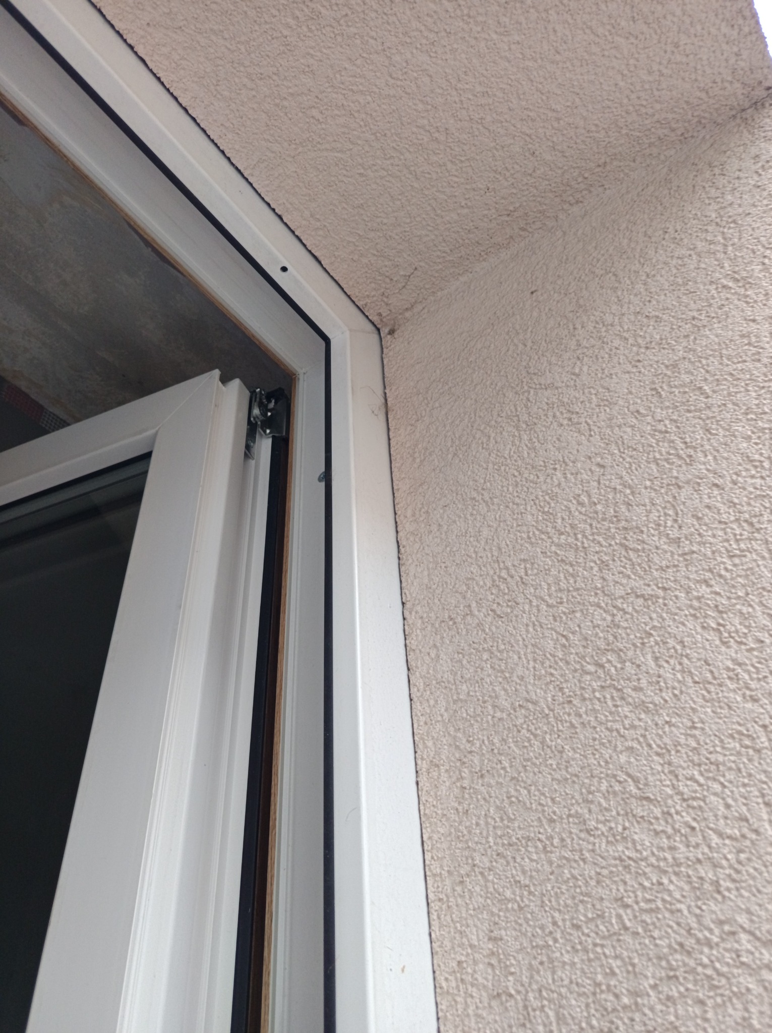 Fragment białej ramy okiennej z widocznym zawiasem, na tle beżowej, chropowatej ściany zewnętrznej. Detal montażu okna.