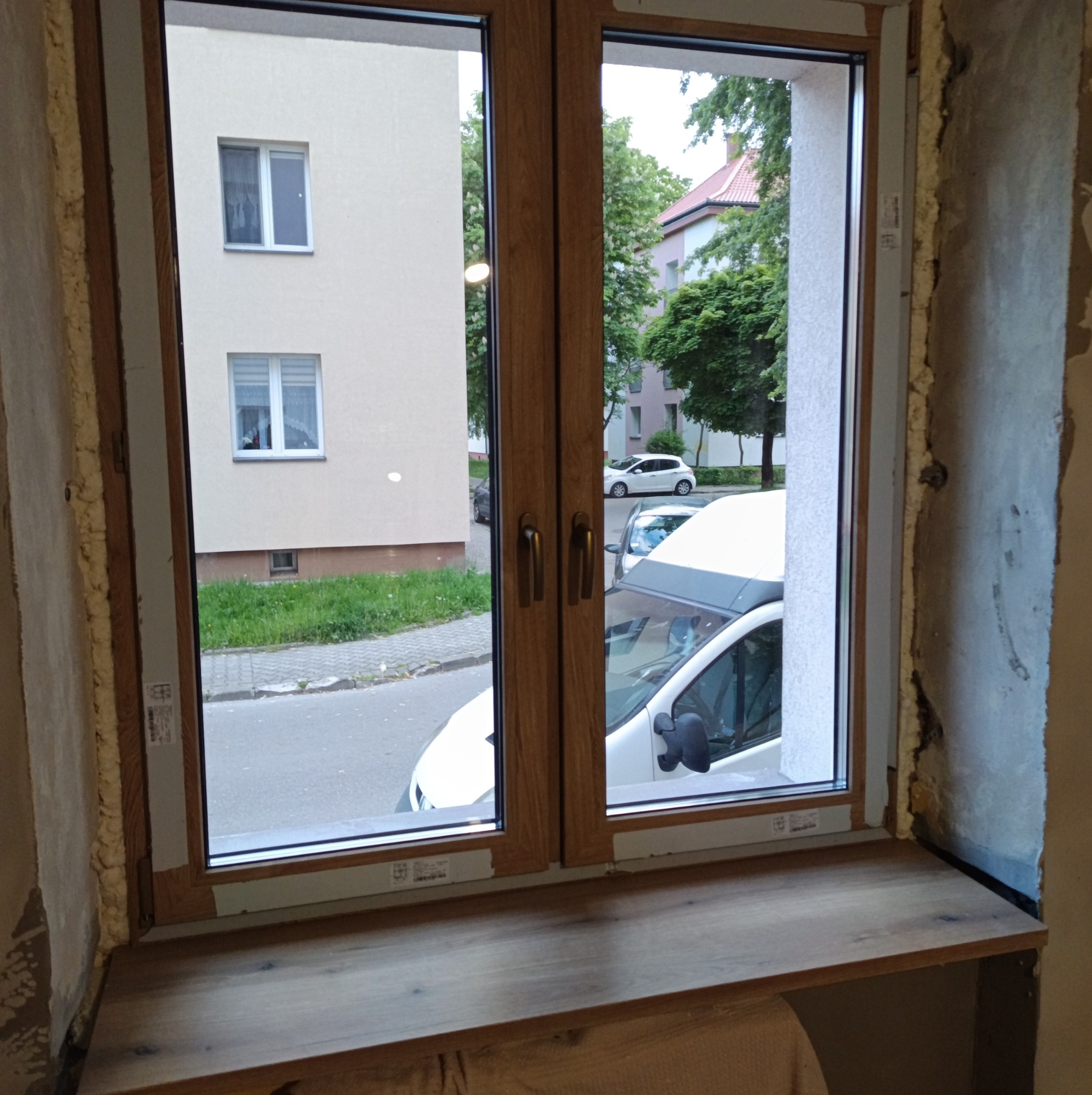 Dwuskrzydłowe okno z drewnianą ramą i parapetem. Widok na ulicę z zaparkowanymi autami. Pianka montażowa wokół ramy. Wnętrze w trakcie wykończenia.