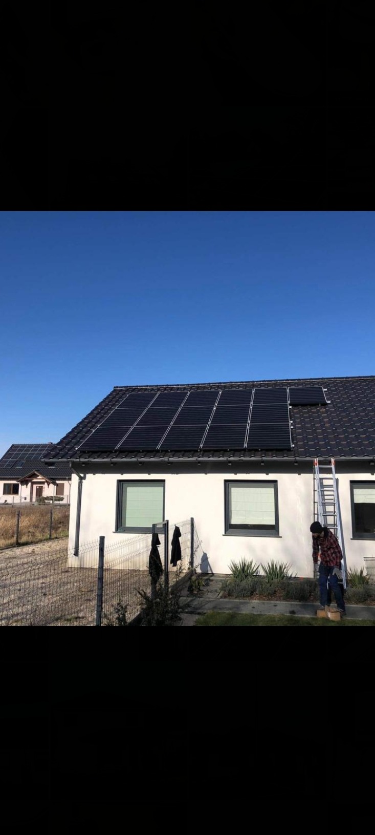 Montaż paneli słonecznych na dachu domu jednorodzinnego; instalator na drabinie w trakcie pracy, widoczne czarne panele na ciemnej dachówce, błękitne niebo w tle.