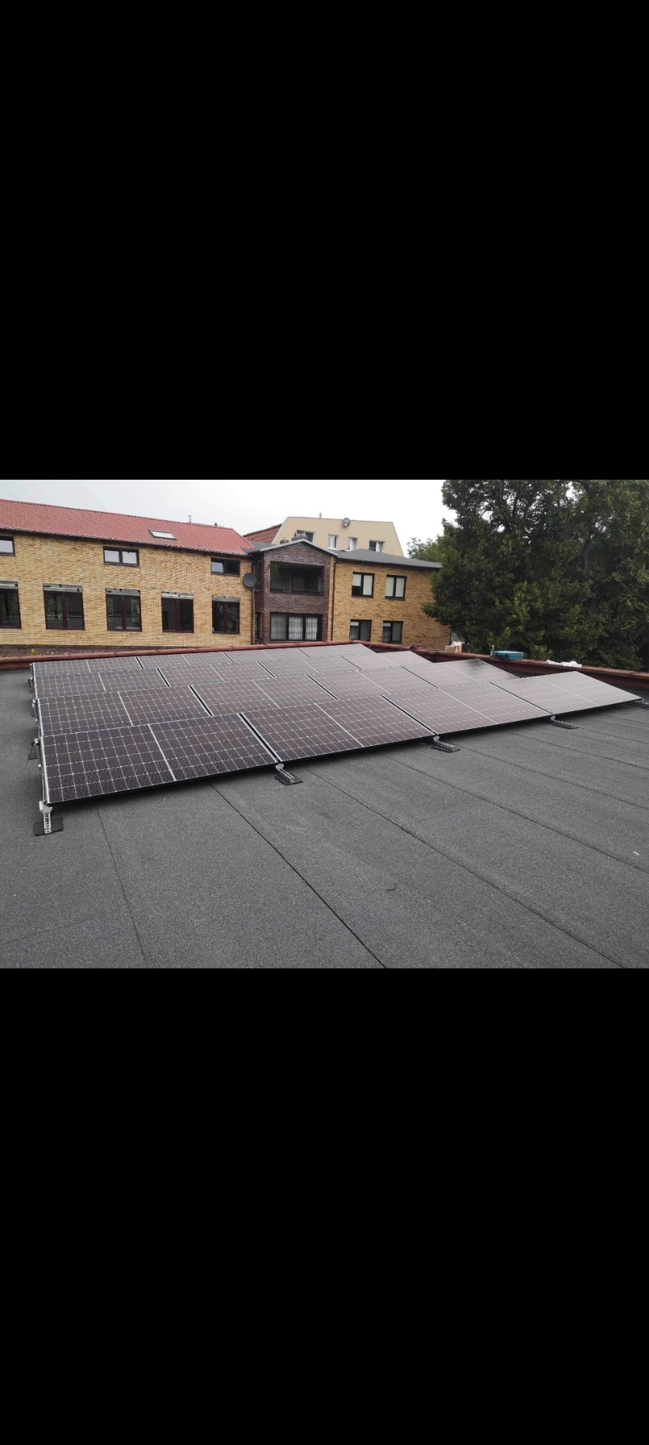 Panele fotowoltaiczne zamontowane na płaskim dachu budynku z żółtej cegły w pochmurny dzień.