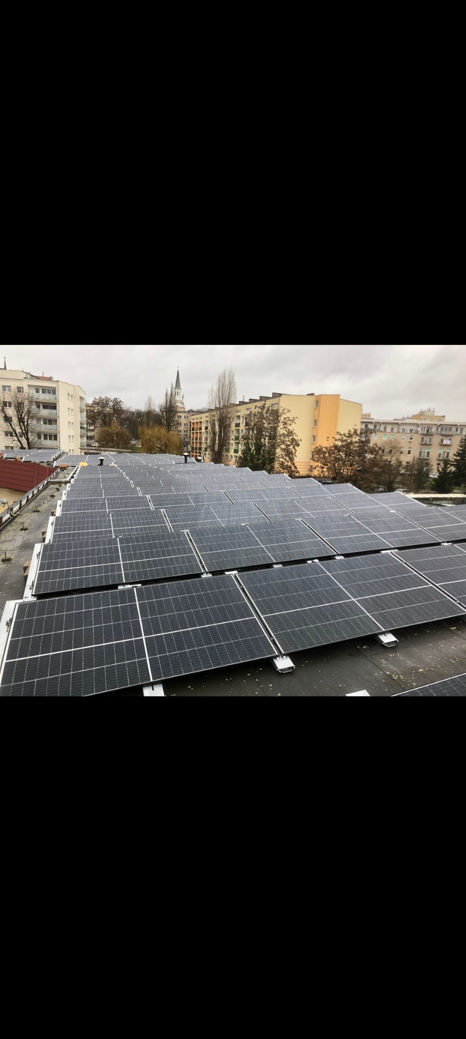 Widok z góry na instalację paneli słonecznych na dachu budynku w pochmurny dzień, w tle budynki mieszkalne i wieża kościelna.