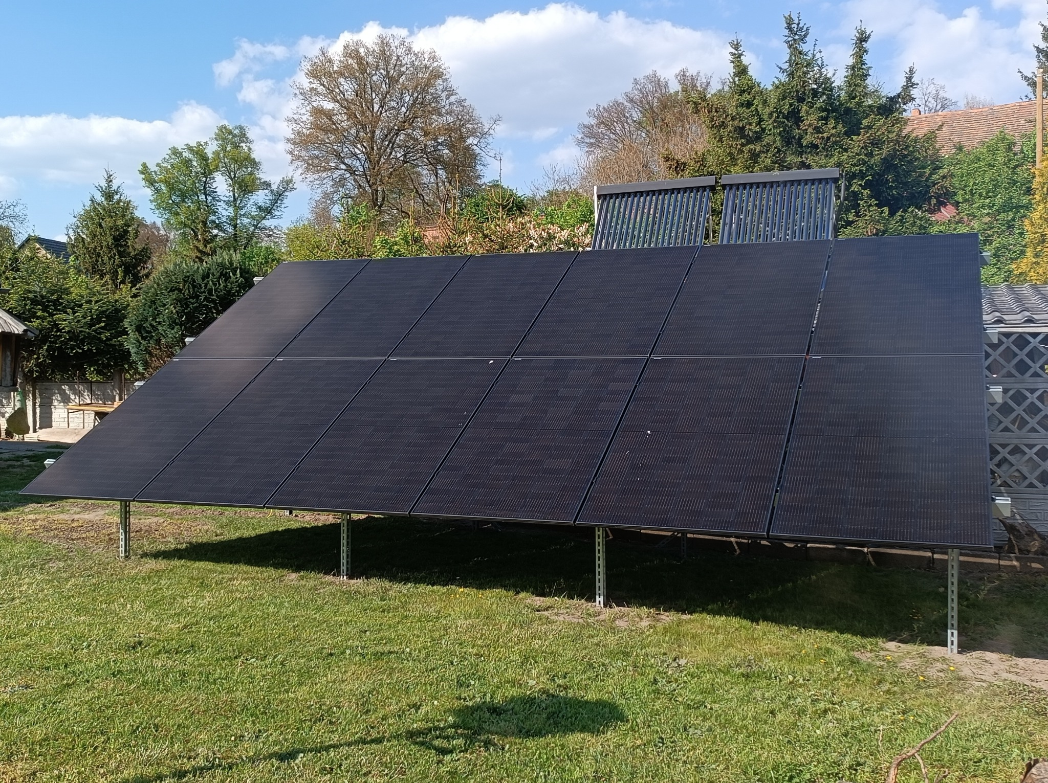 Czarny panel fotowoltaiczny na metalowych wspornikach, ustawiony na trawniku przed domem z kolektorami słonecznymi na dachu w tle, w słoneczny dzień.