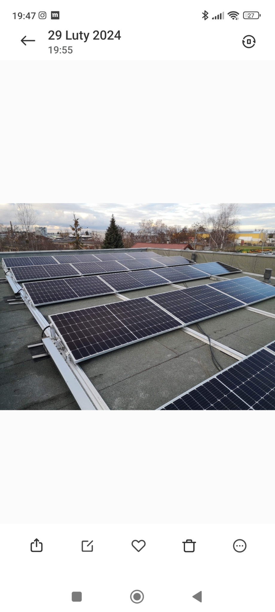 Rząd paneli słonecznych na płaskim dachu budynku, widoczne mocowania i przewody, w tle drzewa i zabudowania miejskie w pochmurny dzień.