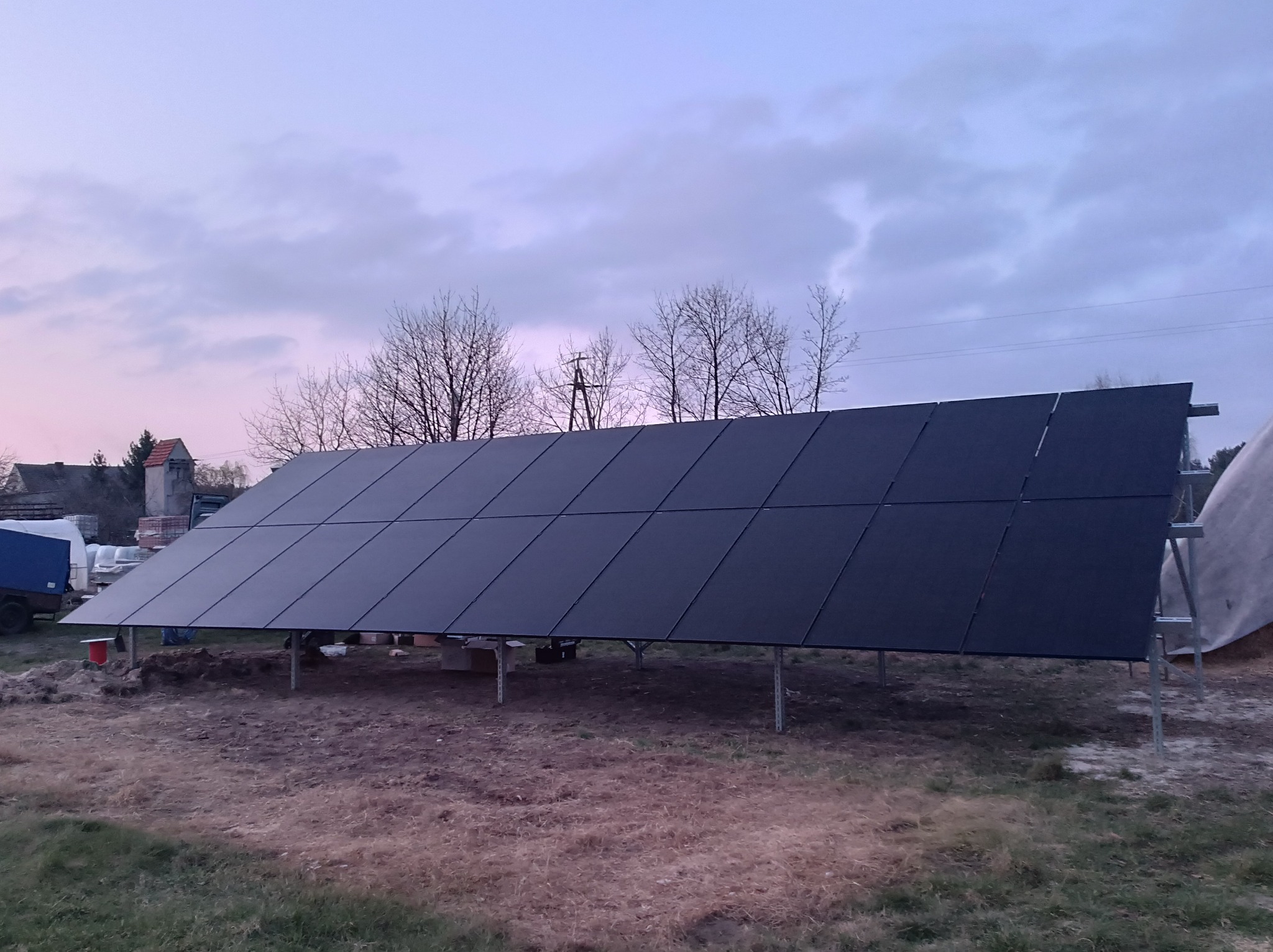 Rząd czarnych paneli słonecznych na metalowej konstrukcji, w tle widoczne drzewa bez liści i budynek z czerwoną wieżyczką, pochmurne niebo.