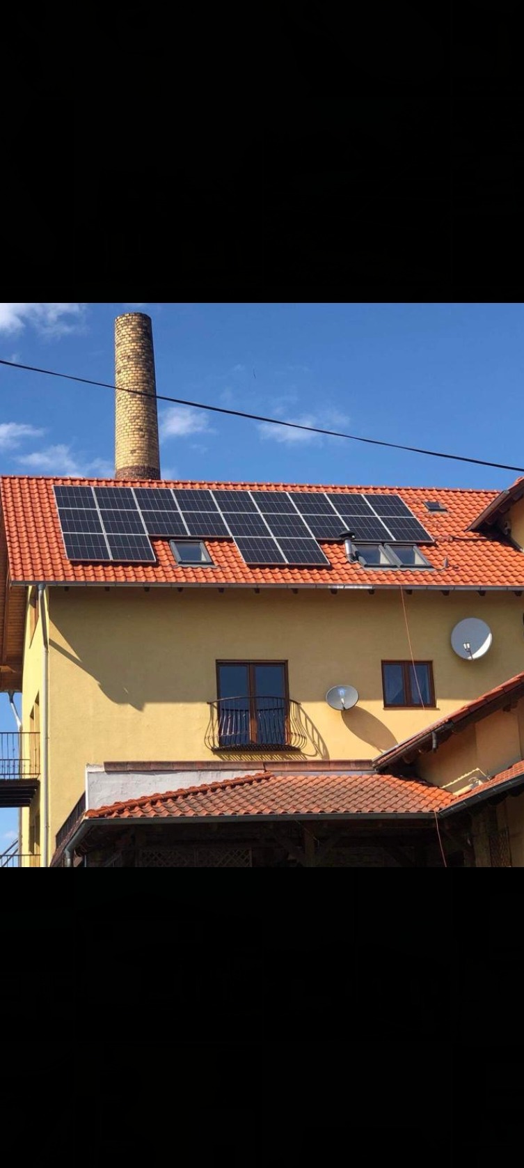 Żółty budynek z pomarańczową dachówką, na której zamontowano panele słoneczne. Na dachu widoczne okna dachowe. W tle komin z cegły i niebieskie niebo.