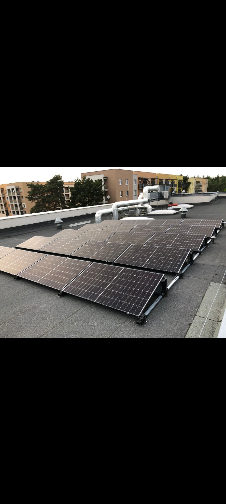 Rząd paneli fotowoltaicznych na płaskim dachu budynku wielorodzinnego, widoczne elementy montażowe i instalacja wentylacyjna w tle.