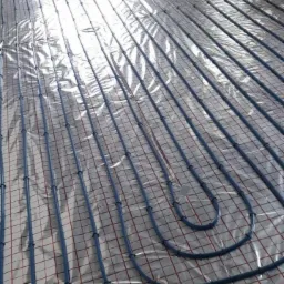 Niskokątowy widok na nieotynkowaną podłogę z rozłożonymi, niebieskimi rurami ogrzewania podłogowego, przymocowanymi do folii odblaskowej z czerwoną siatką.