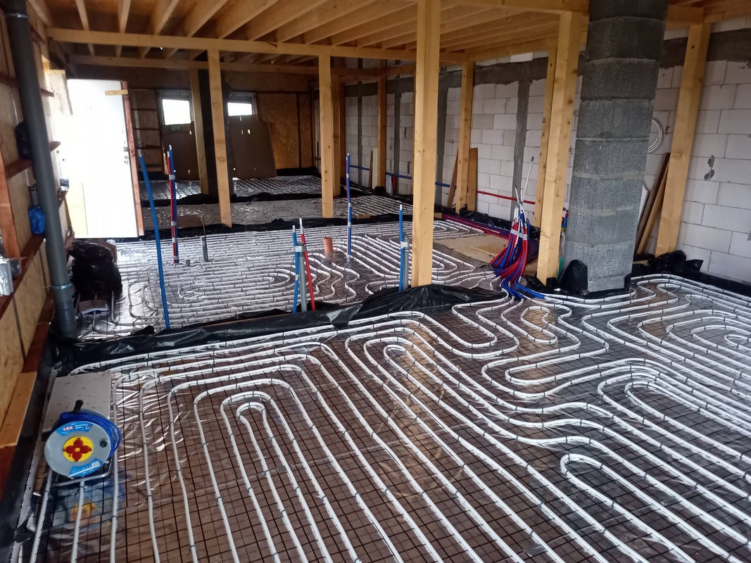 Rozłożone białe rury ogrzewania podłogowego na czarnej folii z widocznymi elementami konstrukcji budynku w surowym stanie.