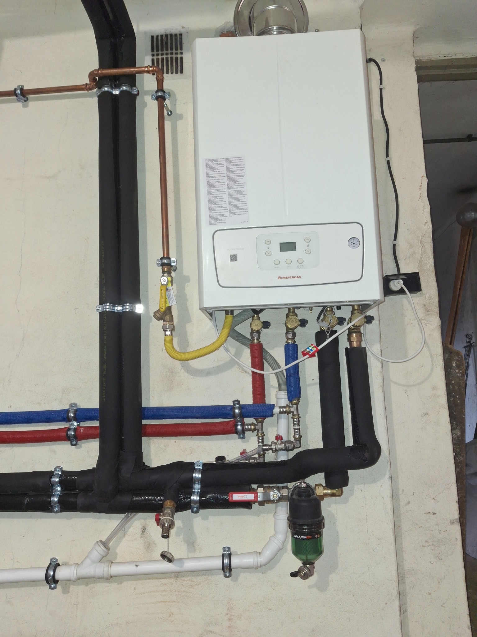 Biały piec gazowy Immergas na ścianie z widocznymi rurami miedzianymi i czarnymi izolowanymi, podłączonymi do instalacji. Żółty przewód gazowy i filtr Valvex na dole.