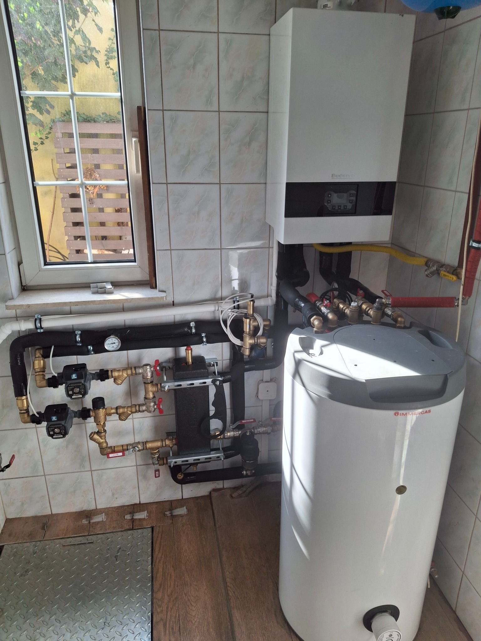 Nowoczesny piec gazowy z zasobnikiem ciepłej wody w kotłowni w Rykach. Widoczne liczne rury i zawory. Okno z widokiem na zewnątrz.