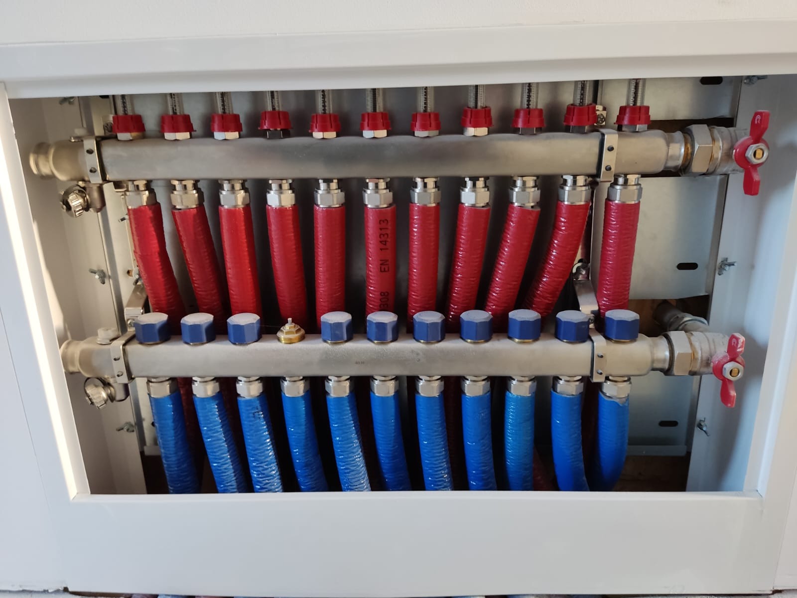 Rozdzielacz instalacji grzewczej podłogowej z czerwonymi i niebieskimi rurami w białej szafce, widoczne zawory i nakrętki.