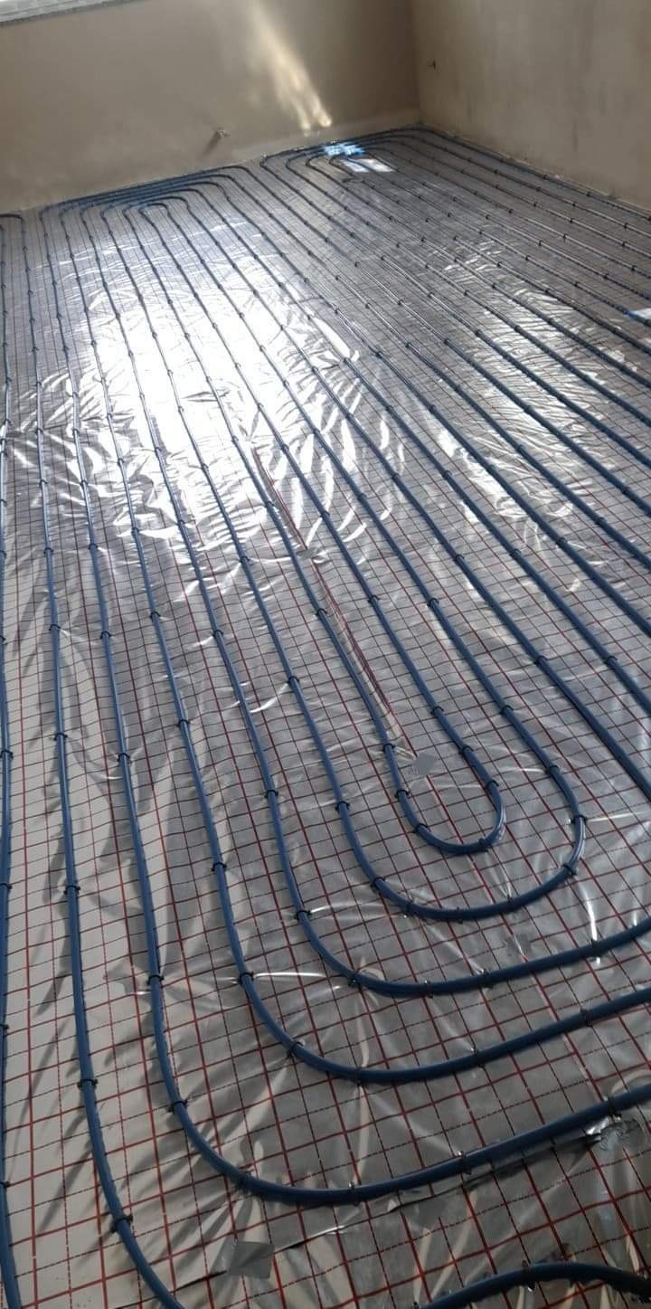 Niskokątowy widok na nieotynkowaną podłogę z rozłożonymi, niebieskimi rurami ogrzewania podłogowego, przymocowanymi do folii odblaskowej z czerwoną siatką.