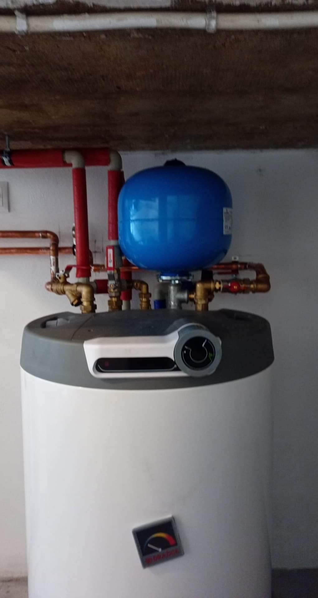 Bojler elektryczny z niebieskim zbiornikiem wyrównawczym, podłączony do instalacji z rur miedzianych i czerwonych, widoczne zawory i manometr, sufit betonowy w tle.