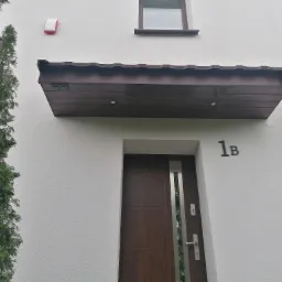 Elewacja budynku z nowoczesnymi drzwiami wejściowymi w kolorze ciemnego drewna, oświetleniem nad drzwiami i numerem domu '1B' w czarnym kolorze, widok z dołu.