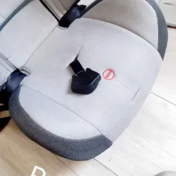 Szare fotelik samochodowy dla dziecka z czarnymi pasami bezpieczeństwa i napisem 'PRESS' na siedzisku, leżący na drewnianej podłodze.