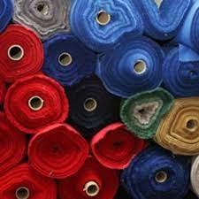 Stos zwiniętych rolek tkanin w różnych kolorach: czerwonym, niebieskim, szarym, czarnym i zielonym. Widoczne rdzenie rolek. Niektóre rolki pogniecione.