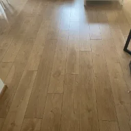 Ułożone panele podłogowe w odcieniach jasnego drewna, widoczne fragmenty białych ścian i nóg krzeseł oraz metalowa podstawa stołka barowego.