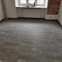 Szare panele podłogowe w pustym pokoju z grzejnikiem pod oknem i fragmentem ściany z cegły