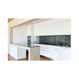 Nowoczesna, minimalistyczna kuchnia z białymi frontami szafek, czarnym blatem i wyspą z betonowym blatem i szklanym przedłużeniem, widok na zabudowany piekarnik.