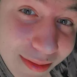 Portret młodej osoby z bliska, z lekkim uśmiechem i widocznym znamieniem na twarzy. Ujęcie z dołu, oświetlenie nierównomierne.