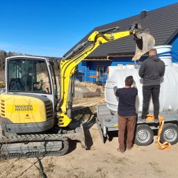 Fundacja Sent - Żółta koparka Neuson umieszcza biały zbiornik na przyczepie, w tle błękitny dom z ciemnym dachem. Dwóch mężczyzn pomaga w załadunku. Słoneczny dzień.