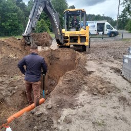 Fundacja Sent - Pracownik z łopatą w wykopie z rurą, koparka Volvo w tle, prace ziemne. Widoczny samochód firmowy z numerem telefonu. Dzień pochmurny.