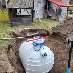 Fundacja Sent - Montaż przydomowej oczyszczalni: biały zbiornik w wykopie, poziomica na górze, rury podłączeniowe. W tle budynek z drewnianą altaną i schodami.