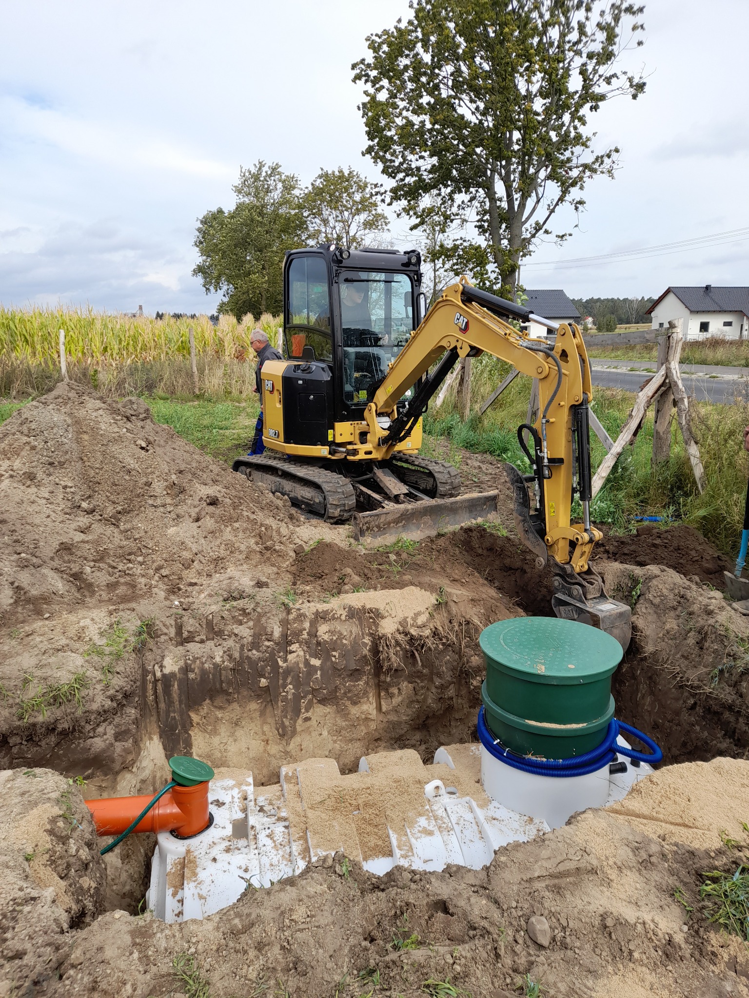 Żółta mini koparka CAT podczas instalacji zbiornika oczyszczalni ścieków w wykopie. Widoczne elementy instalacji: biały zbiornik z zielonymi pokrywami, pomarańczowa rura. W tle pole kukurydzy...