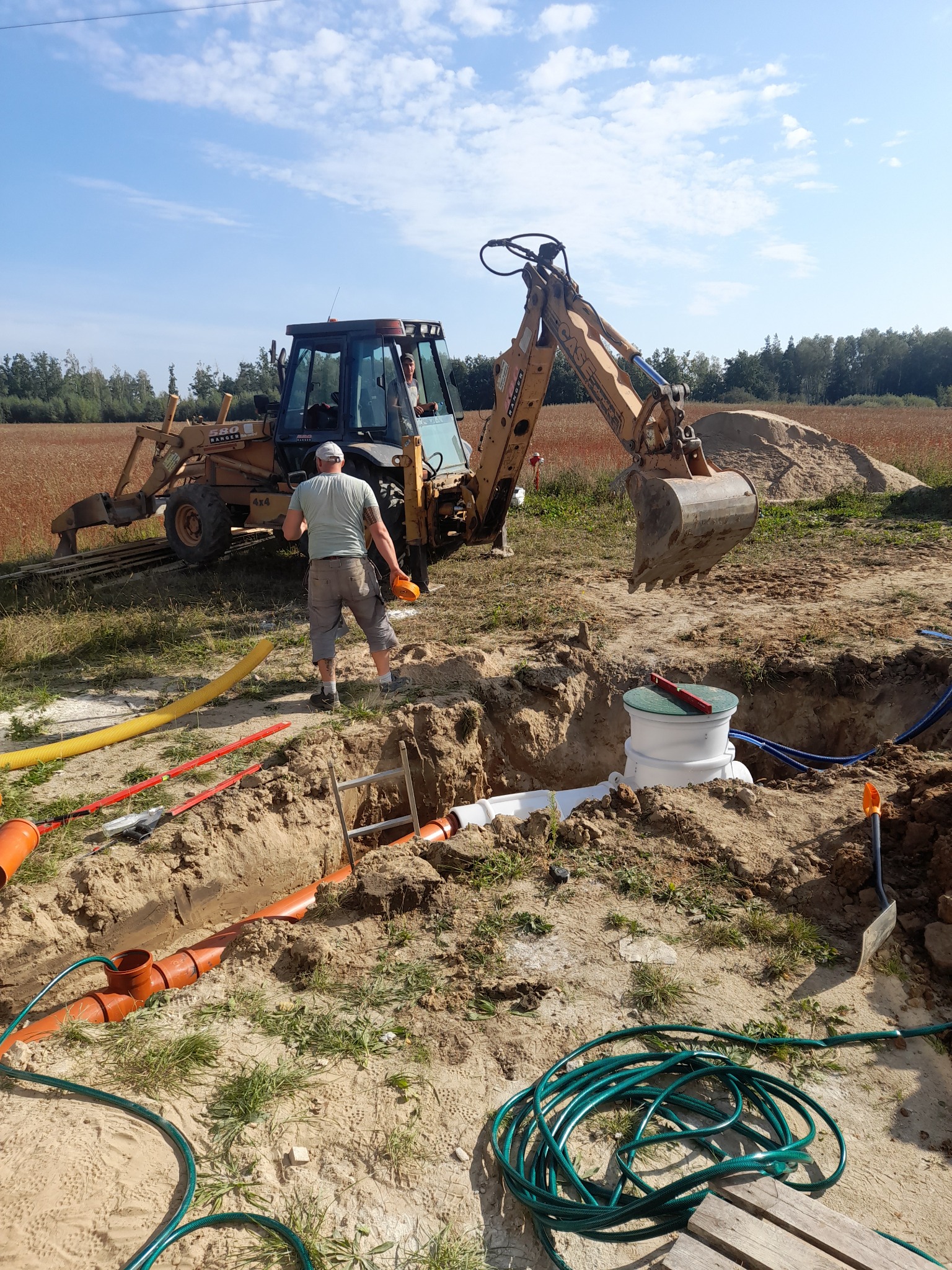 Montaż podziemnego zbiornika z rurami, widoczny wykop, poziomica na pokrywie, koparka w tle i pracownik z miarką.