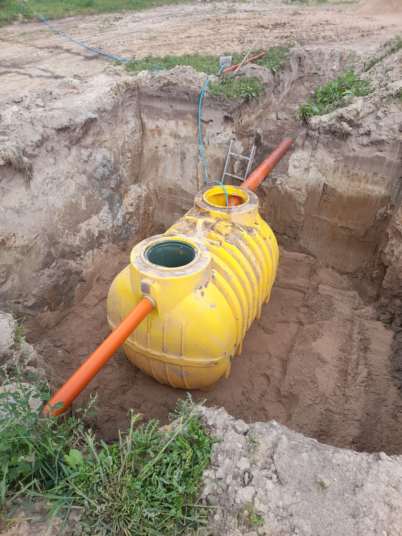 Żółty zbiornik z rurami w wykopie, widoczne ślady ponownego montażu, gliniasta gleba, drabina oparta o ścianę wykopu.