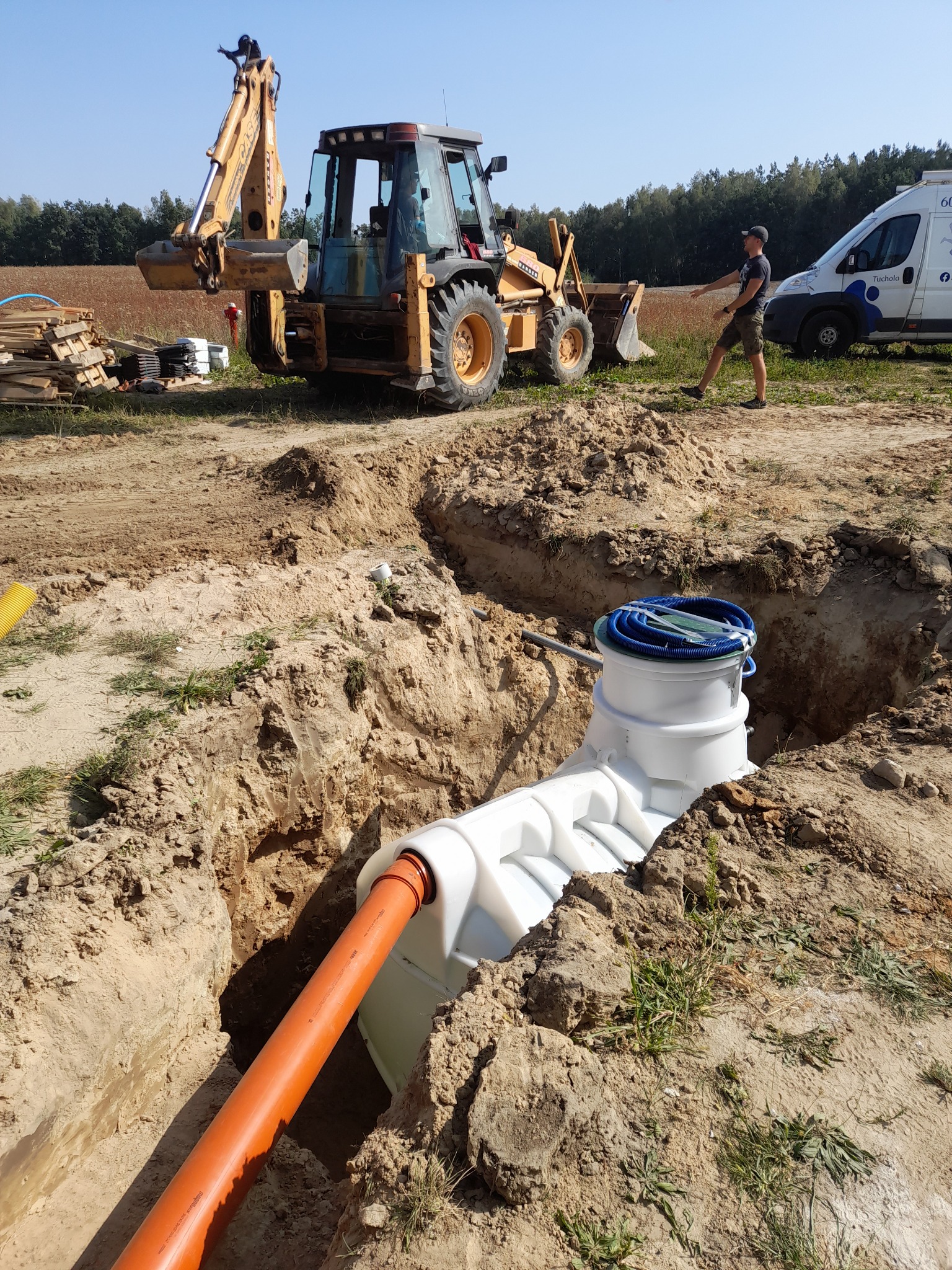 Montaż zbiornika biologicznego w wykopie, widoczna pomarańczowa rura doprowadzająca ścieki, biały zbiornik z niebieskimi przewodami na górze, w tle koparka Case, palety z materiałami, bus firmowy...