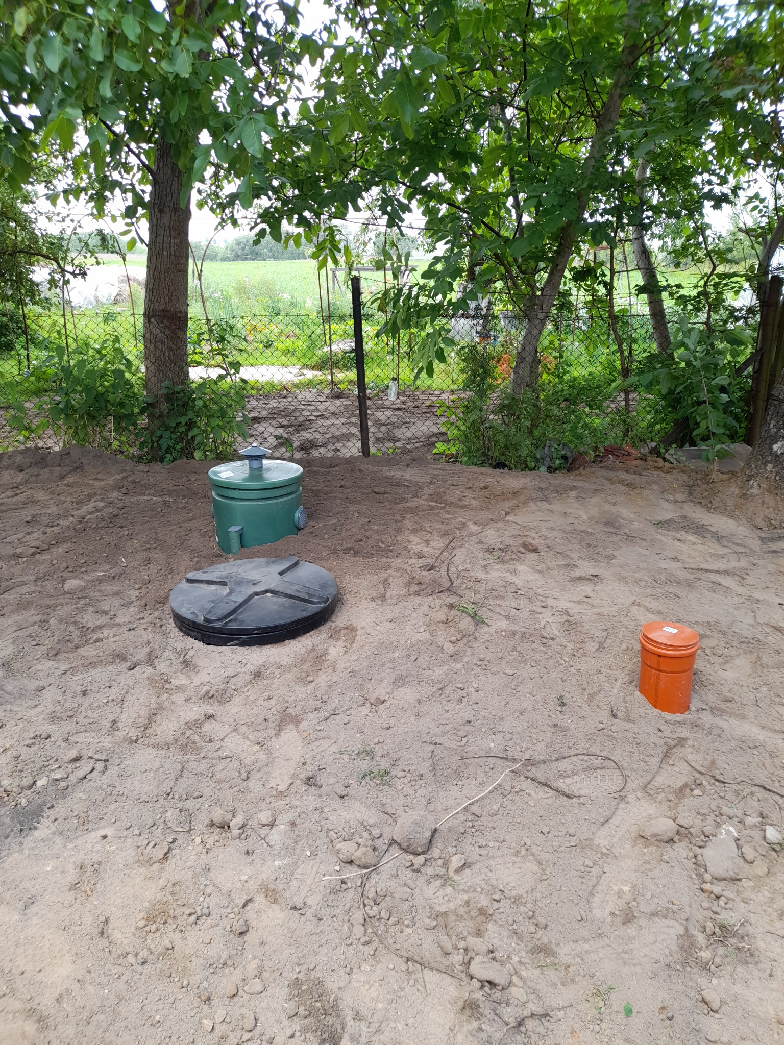 Zielony zbiornik i czarna pokrywa oczyszczalni ścieków na piaszczystym podłożu w Tucholi. W tle zieleń drzew i krzewów, ogrodzenie z siatki.