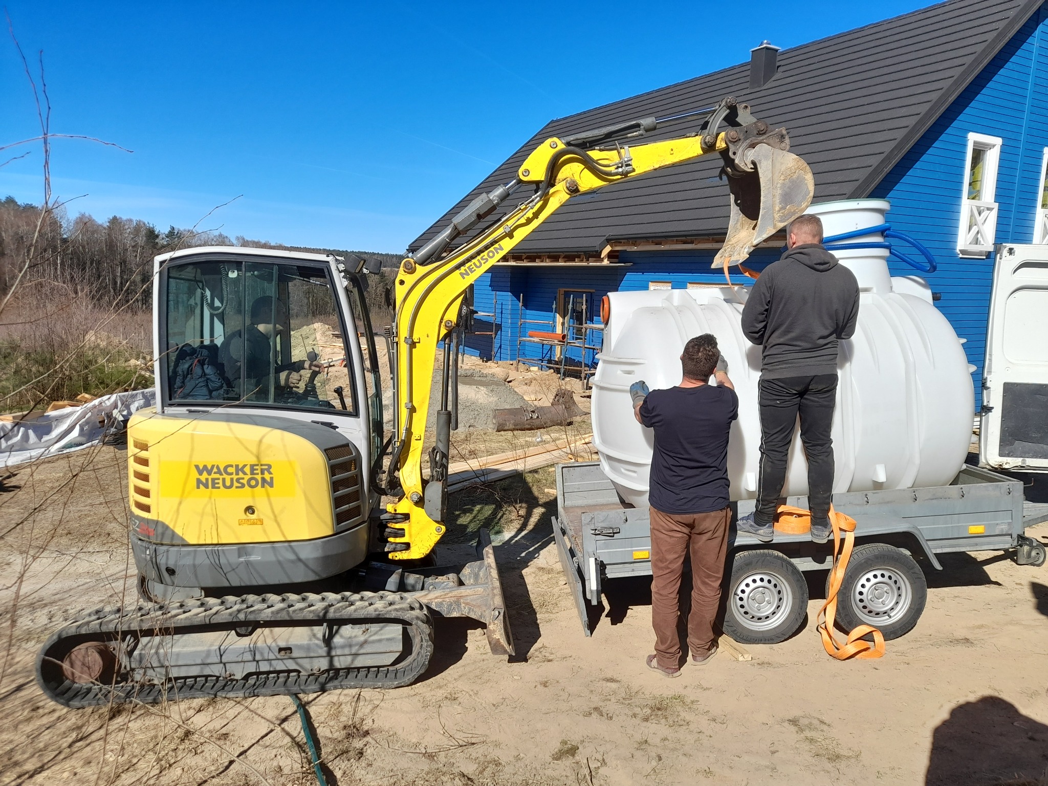 Żółta koparka Neuson umieszcza biały zbiornik na przyczepie, w tle błękitny dom z ciemnym dachem. Dwóch mężczyzn pomaga w załadunku. Słoneczny dzień.