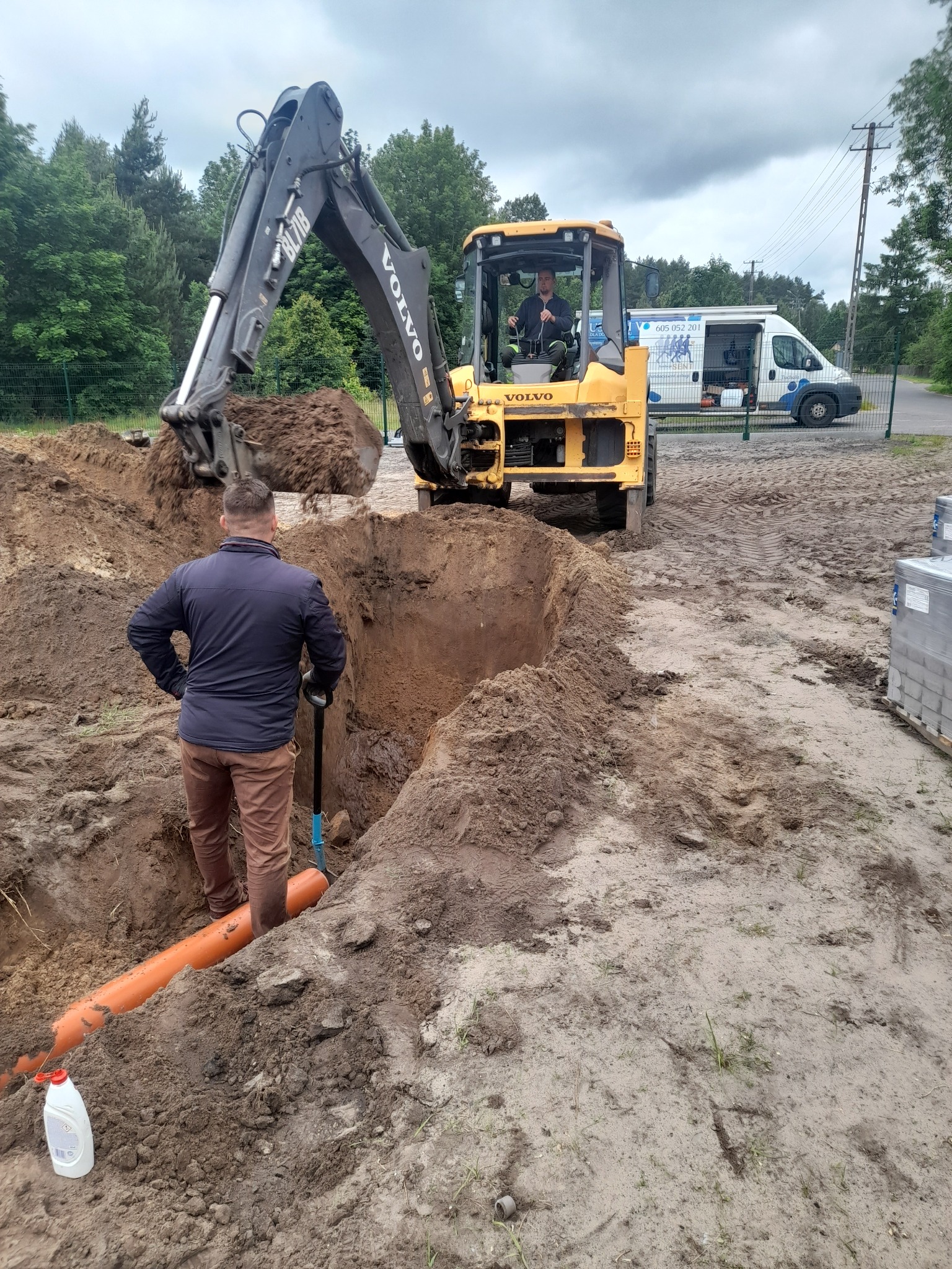 Pracownik z łopatą w wykopie z rurą, koparka Volvo w tle, prace ziemne. Widoczny samochód firmowy z numerem telefonu. Dzień pochmurny.