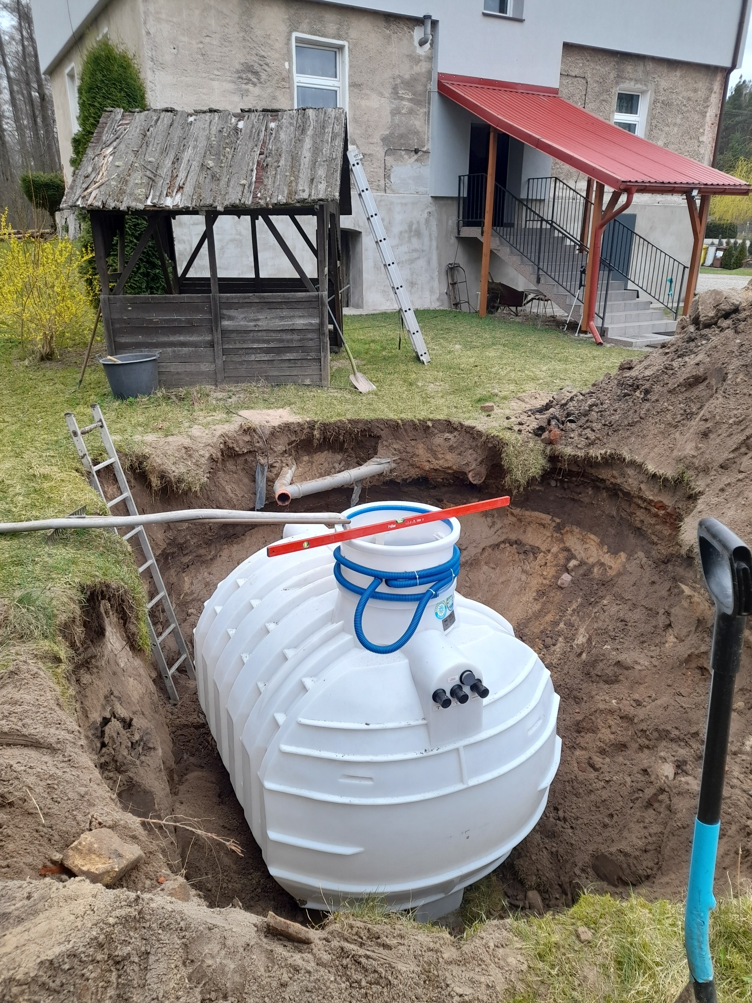 Montaż przydomowej oczyszczalni: biały zbiornik w wykopie, poziomica na górze, rury podłączeniowe. W tle budynek z drewnianą altaną i schodami.