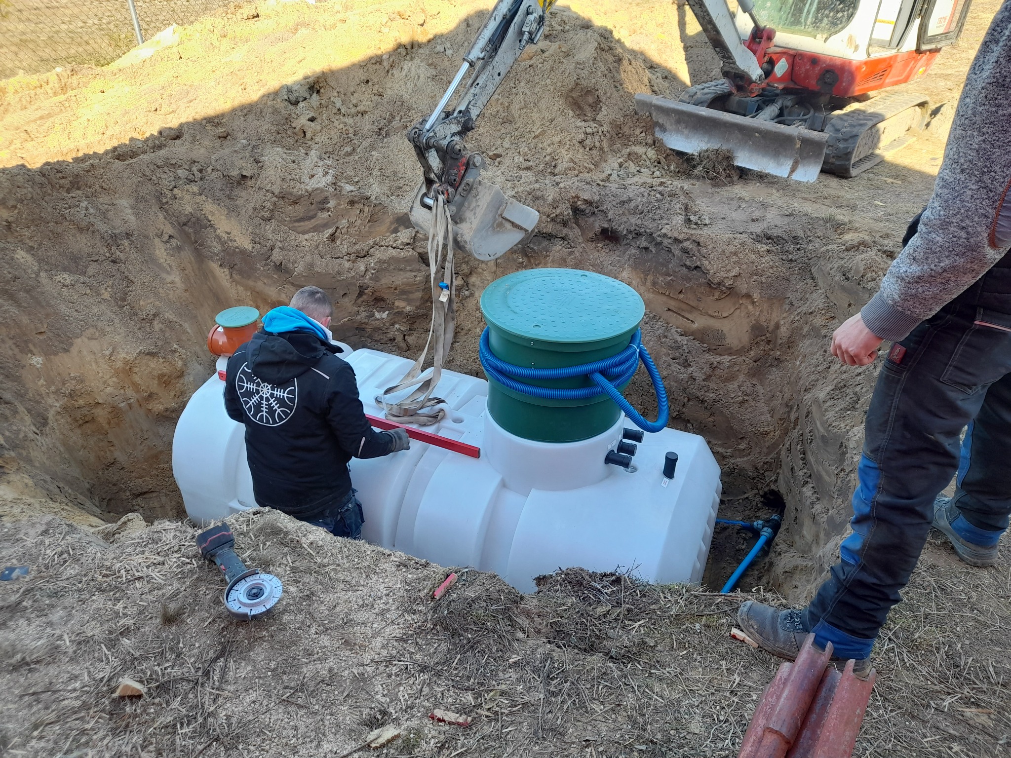 Montaż przydomowej oczyszczalni: pracownik z poziomicą obok zbiornika w wykopie, koparka w tle. Widoczne narzędzia i rury. Dzień, ziemia, instalacja.