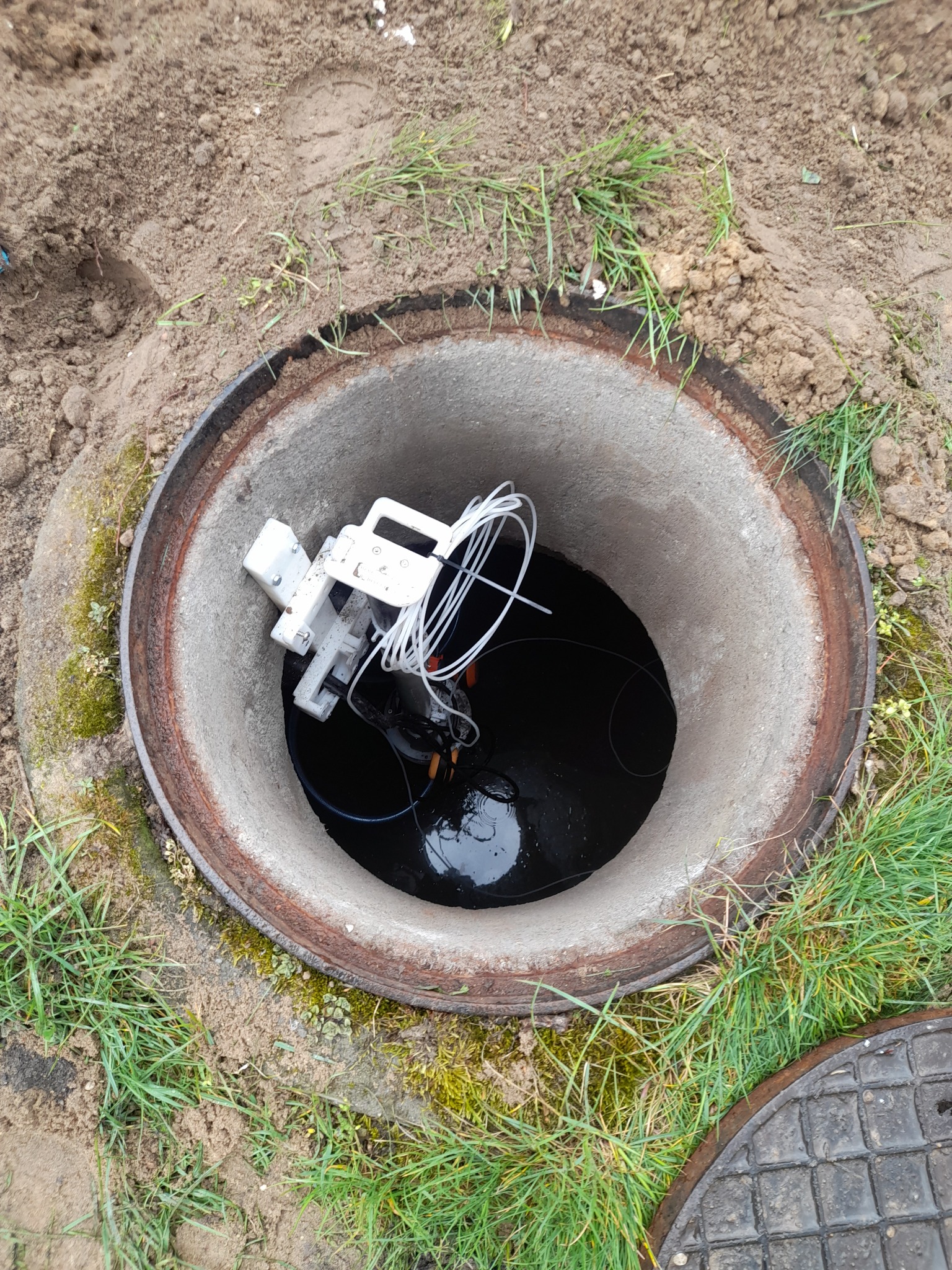 Betonowy krąg studni z wodą i zanurzonym urządzeniem pomiarowym, widok z góry. Wokół ziemia i trawa.