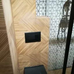 Nowoczesna łazienka z geometrycznymi płytkami drewnopodobnymi na ścianach, czarnym przyciskiem spłuczki i czarną miską WC, kabina prysznicowa z heksagonalnymi płytkami w tle.