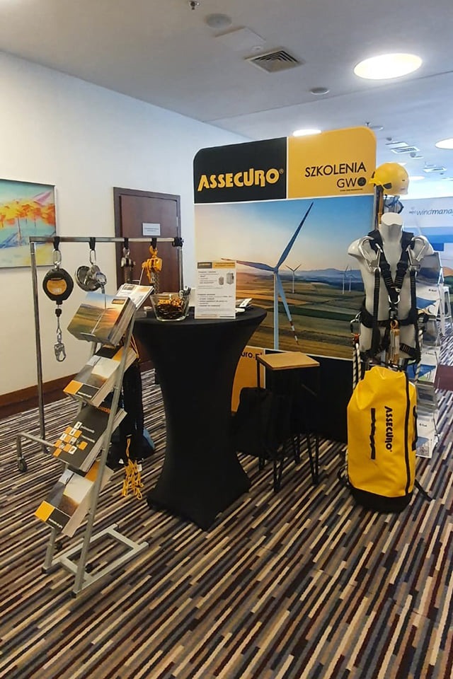 Ekspozycja sprzętu asekuracyjnego firmy Assecuro na tle grafiki z wiatrakami, obejmująca uprząż na manekinie, torbę, zawiesia i karabinki na stojaku oraz ulotki informacyjne.