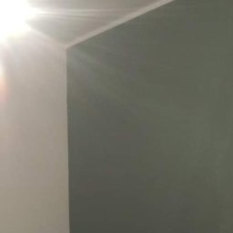 RemoPasja - Świeżo pomalowany narożnik pokoju, gdzie jedna ściana jest biała, a druga ma odcień szarości, widoczne delikatne smugi światła.