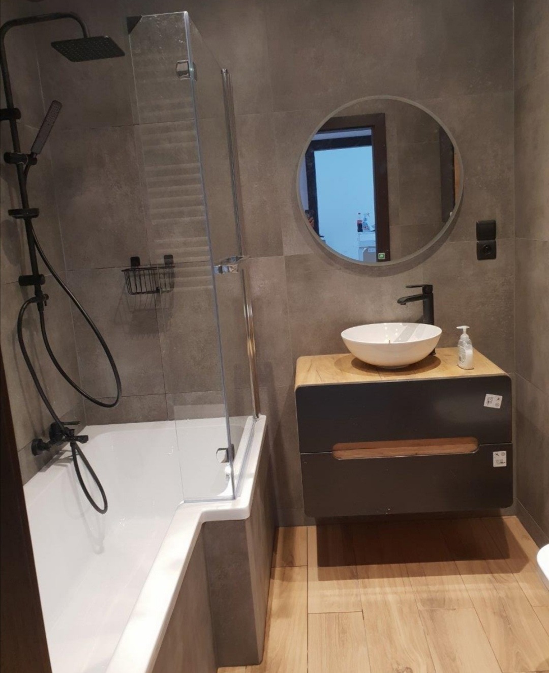 Nowoczesna łazienka w szarościach z drewnianymi akcentami: biała wanna z czarną armaturą i szklanym parawanem, okrągłe lustro nad umywalką nablatową na szafce z drewnianym blatem, drewniana podłoga.