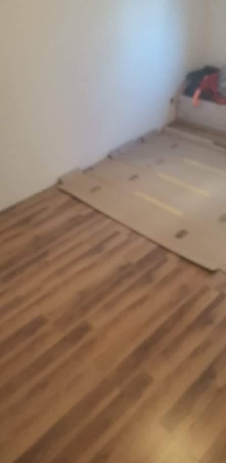 Fragment podłogi z ułożonymi panelami, częściowo zakrytymi kartonem ochronnym, widoczne łączenia i wzór drewna.