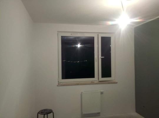 Świeżo odmalowane wnętrze pokoju z białymi ścianami, białym oknem, grzejnikiem i widokiem na nocne miasto, z fragmentem szarej ściany po prawej stronie i małym stołkiem w rogu.