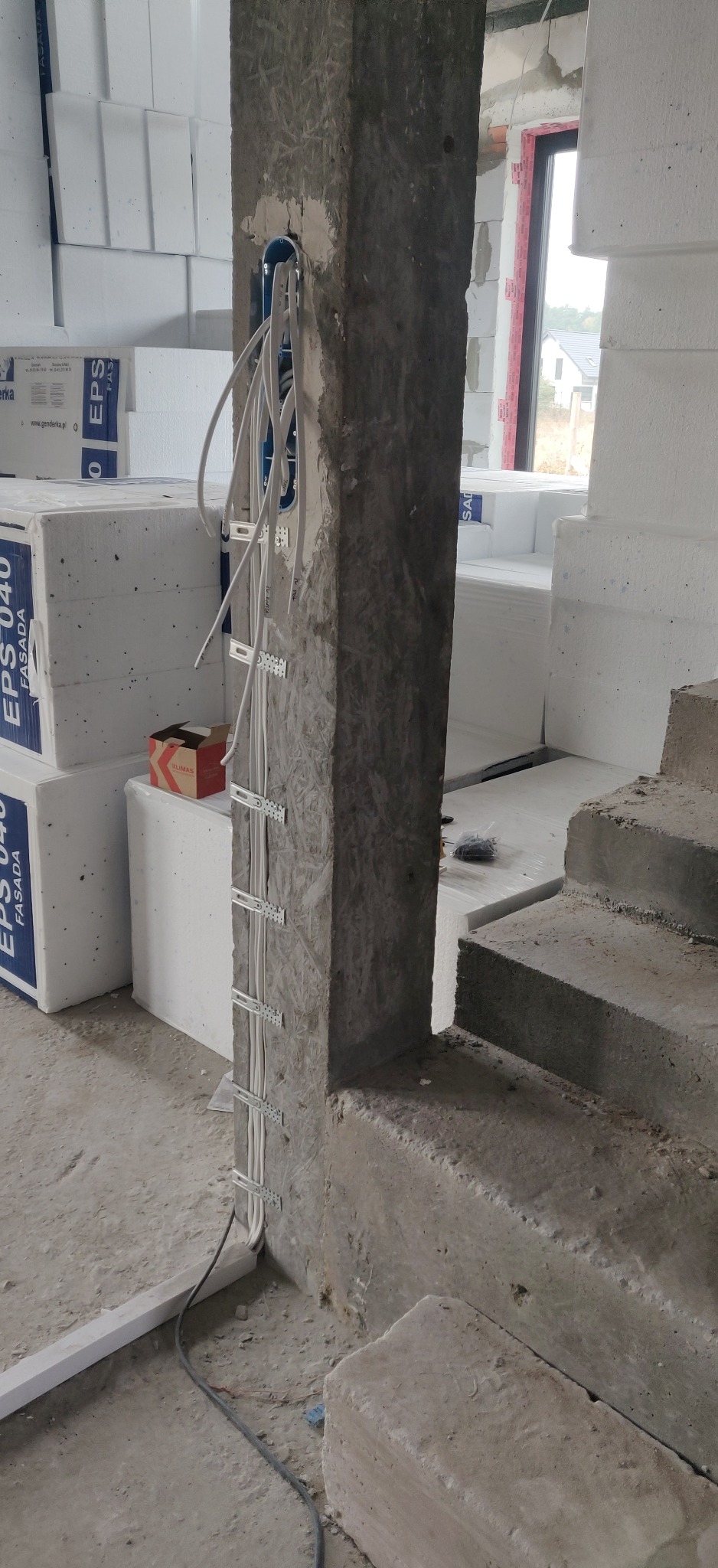 Wiązka przewodów elektrycznych przymocowana metalowymi uchwytami do surowego słupa konstrukcyjnego w trakcie budowy domu jednorodzinnego, widoczne puszki instalacyjne i materiały izolacyjne.