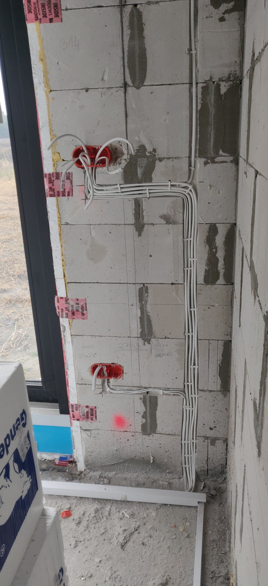 Wewnętrzna ściana z betonu komórkowego w trakcie układania instalacji elektrycznej: widoczne kable prowadzone w peszlach, puszki instalacyjne w kolorze czerwonym, oznaczenia na ścianie. Częściowo...