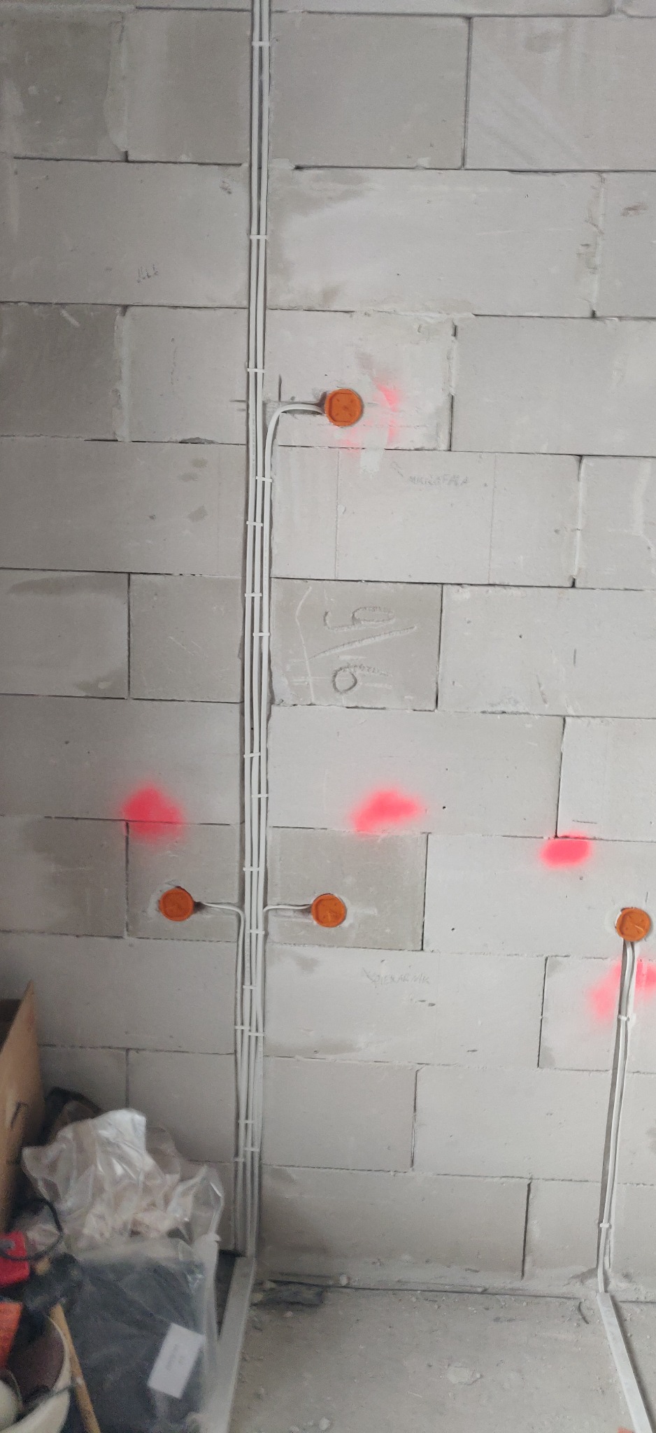 Ściana z betonu komórkowego z zamontowanymi, białymi rurkami instalacyjnymi i pomarańczowymi puszkami elektrycznymi, oznaczenia natryskową farbą w kolorze czerwonym, widoczne przewody.