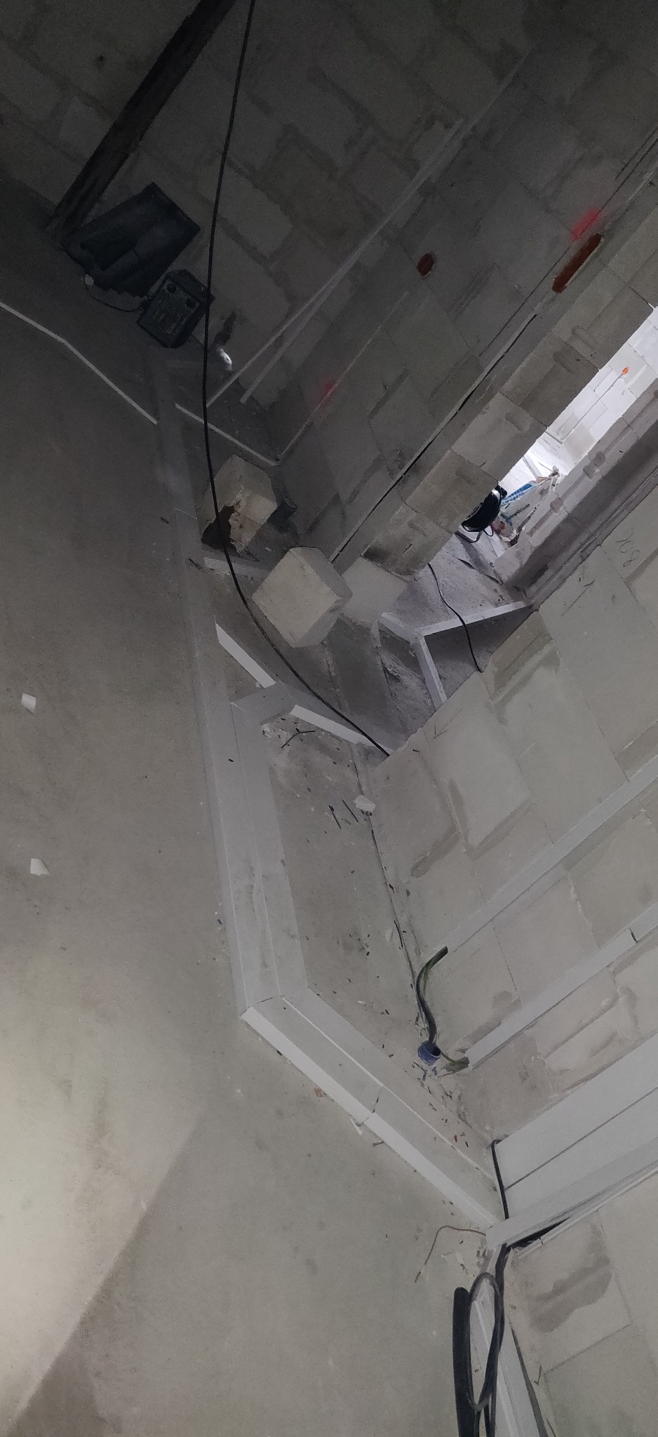 Surowy stan wnętrza domu w trakcie prac instalacyjnych, widoczne kable elektryczne wystające ze ściany i sufitu, białe listwy przypodłogowe, ściany w stanie deweloperskim, betonowy sufit...