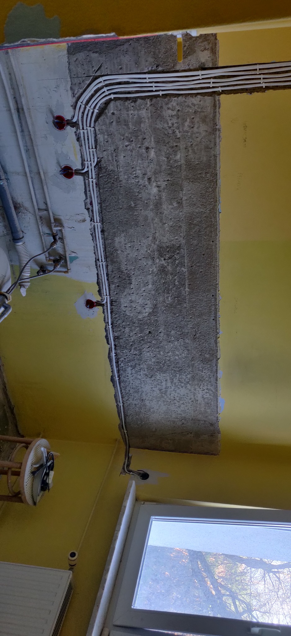 Żółte wnętrze w trakcie remontu, widoczne nowe przewody elektryczne w białych peszlach, mocowane do surowego betonowego stropu, z widocznymi puszkami instalacyjnymi.