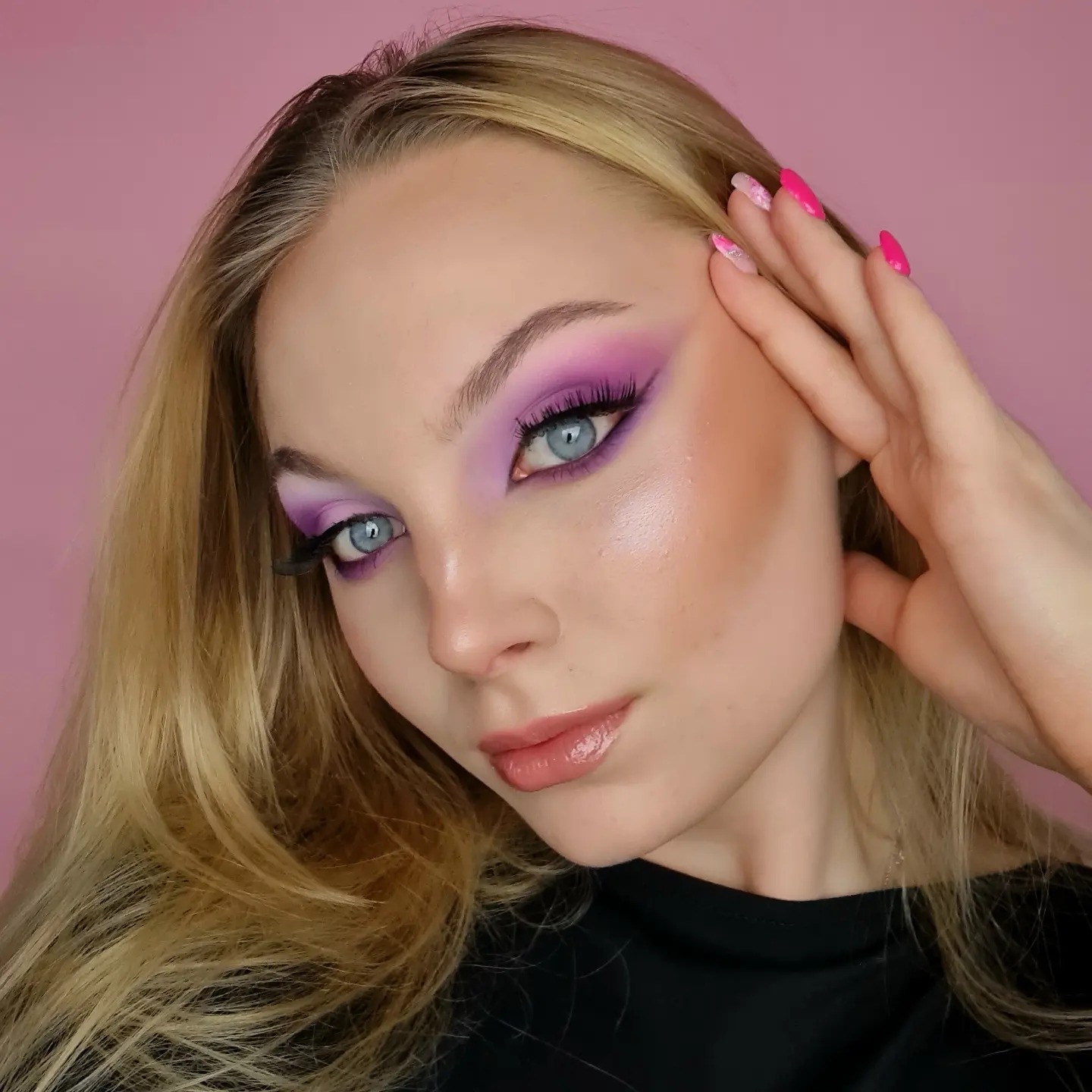 Portret młodej kobiety z intensywnym fioletowo-różowym cieniowaniem powiek, sztucznymi rzęsami i błyszczącymi ustami, pozującej na tle różowej ściany, z dłonią przy twarzy, eksponując manicure...
