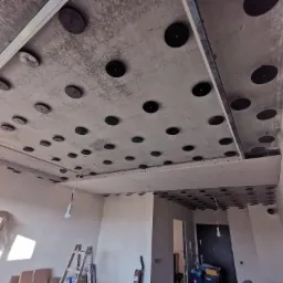 Poddasze w trakcie ocieplania, widoczne czarne okrągłe mocowania na suficie betonowym, metalowe profile oraz częściowo zamontowane płyty gipsowo-kartonowe, drabina oparta o ścianę.